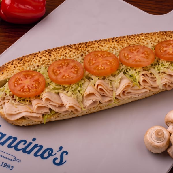 Sesame-seed sub sandwich layered with folded deli meat, shredded lettuce, and tomato slices on a Mancino’s serving board.