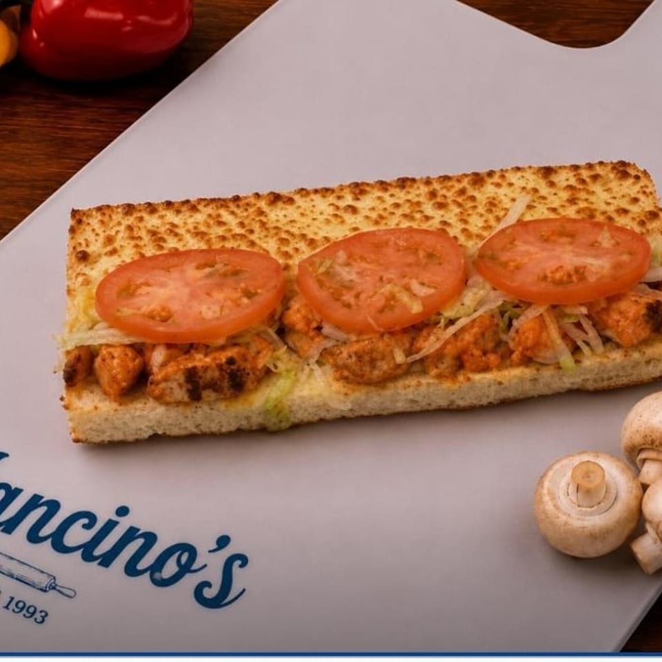 Toasted chicken sandwich with lettuce and sliced tomatoes on a Mancino’s cutting board, surrounded by peppers and mushrooms.