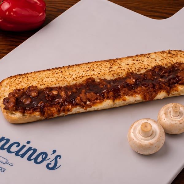Long toasted sandwich filled with saucy chopped meat on a Mancino’s branded plate, garnished with mushrooms and surrounded by peppers.