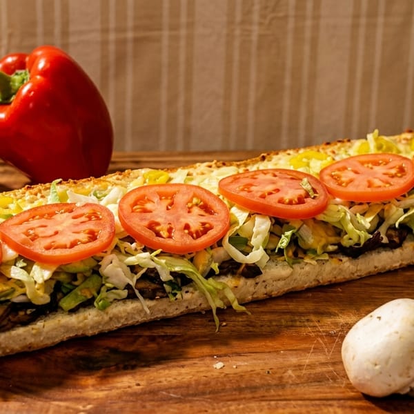 A long submarine sandwich with tomatoes, lettuce, and mushrooms, next to bell peppers and mushrooms.