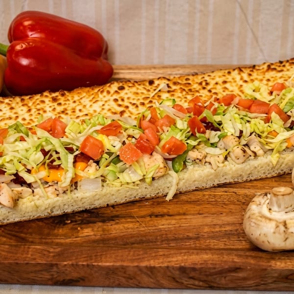 A large chicken sub sandwich with lettuce, tomatoes, and cheese on a wooden board with peppers and mushrooms.